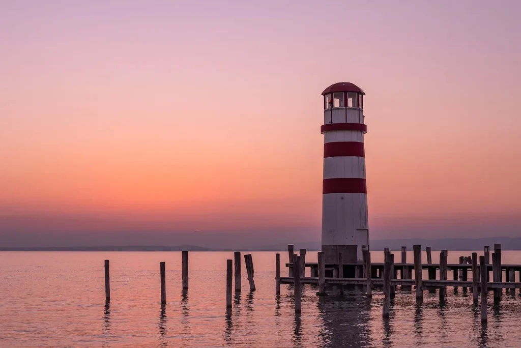 Red and white striped lighthouse on a wooden pier over calm water at sunset