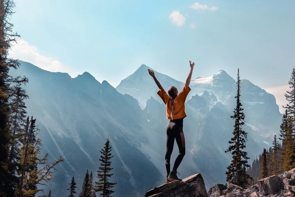 Woman in an orange shirt standing on a rock with arms raised toward a snow-dusted mountain range